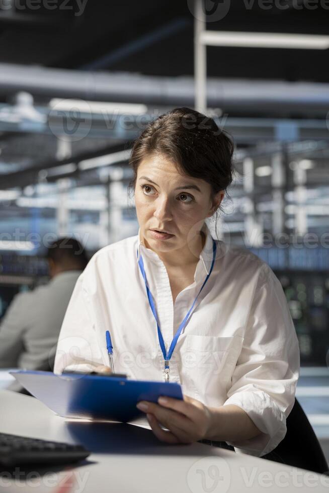 Server room system administrator reviewing documentation on clipboard ahead of scheduled audit. Data center admin verifying configuration data and compliance standards at desk photo