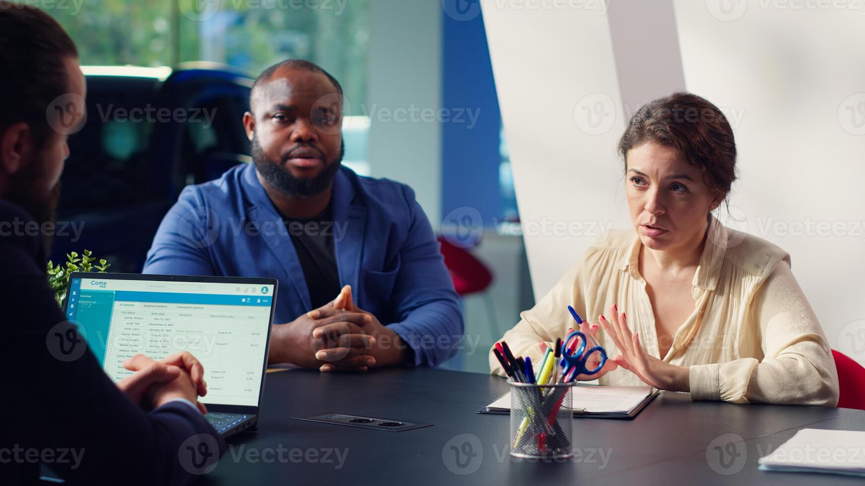 Customers in showroom completing credit application after being sold car insurance by salesman. Clients in dealership signing financing paperwork after buying extra vehicle warranty, camera A photo