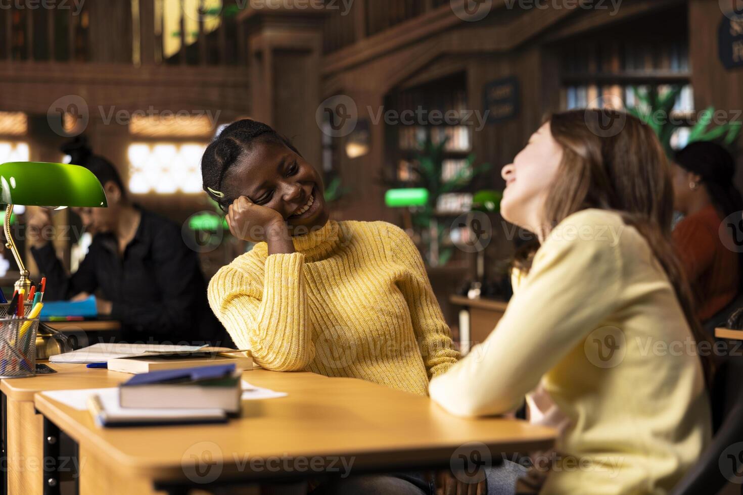Cute teens friends gossiping together and laughing in the library, taking a break from school work and enjoying team project with each other. Young girls enjoying a cheerful conversation. photo