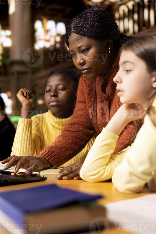 African american professor mentoring diverse students in the library, learning together in an academic library filled with curriculum books. Woman tutoring her class pupils on literature subject. photo