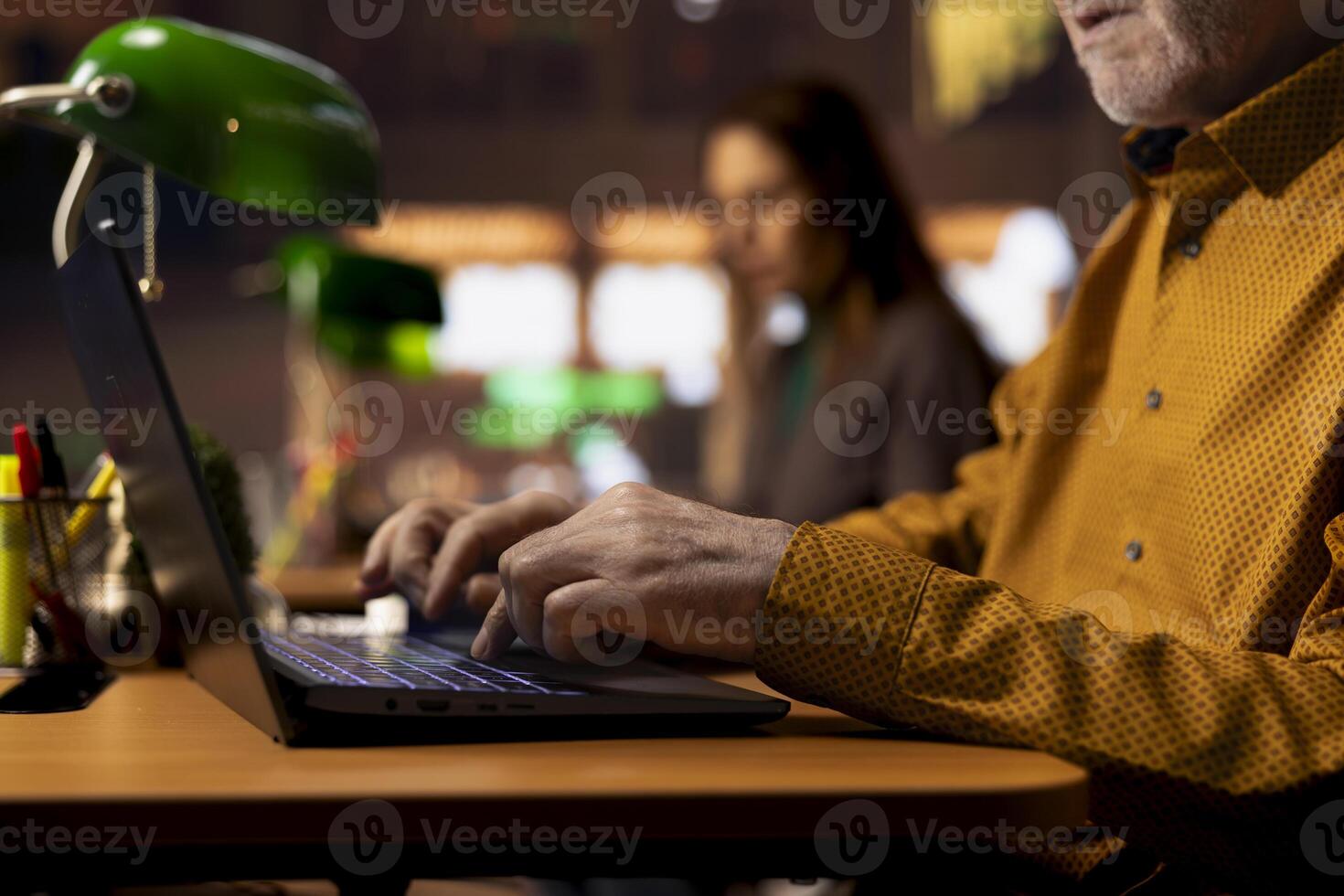 Aged man in a public library preparing thesis paperwork under a green lamp, surrounded by textbooks and study materials. Old pupil reflecting ambition for learning and achievements. photo