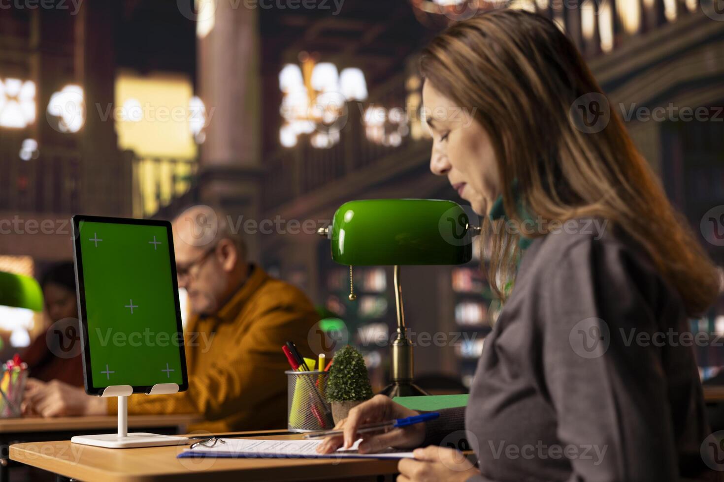 Mature female student reading and writing at desk next to green screen, studying documents and doing her research to use important citations in her master degree thesis. Bookworm. photo