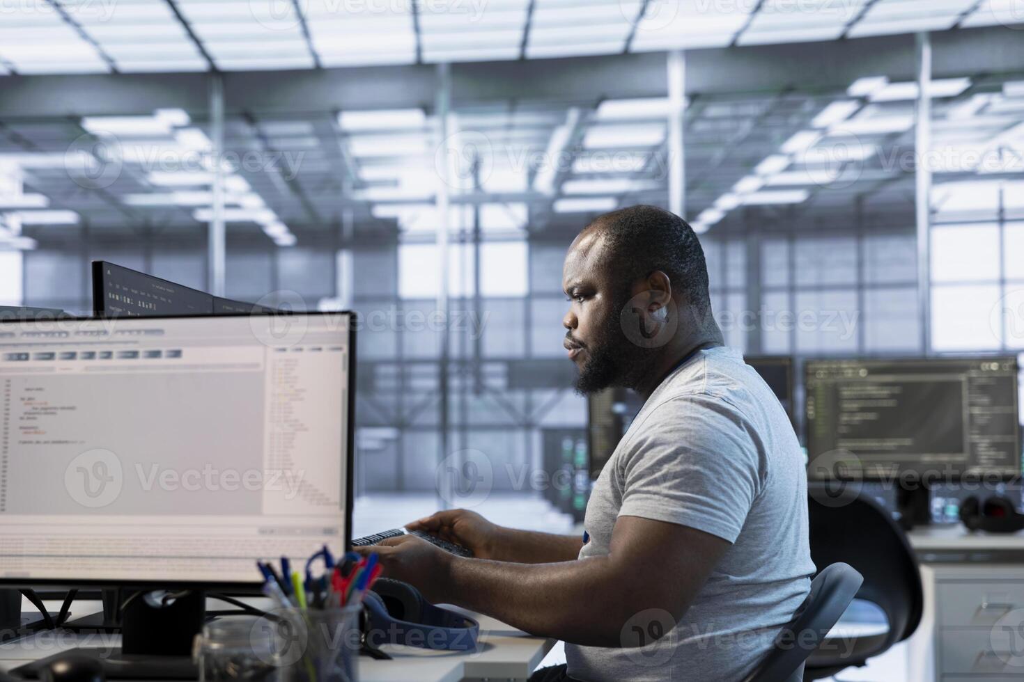Technician at desk writing code and doing checkup on server room providing computing resources for different workloads. African american man investigating data center mainframes using computer photo