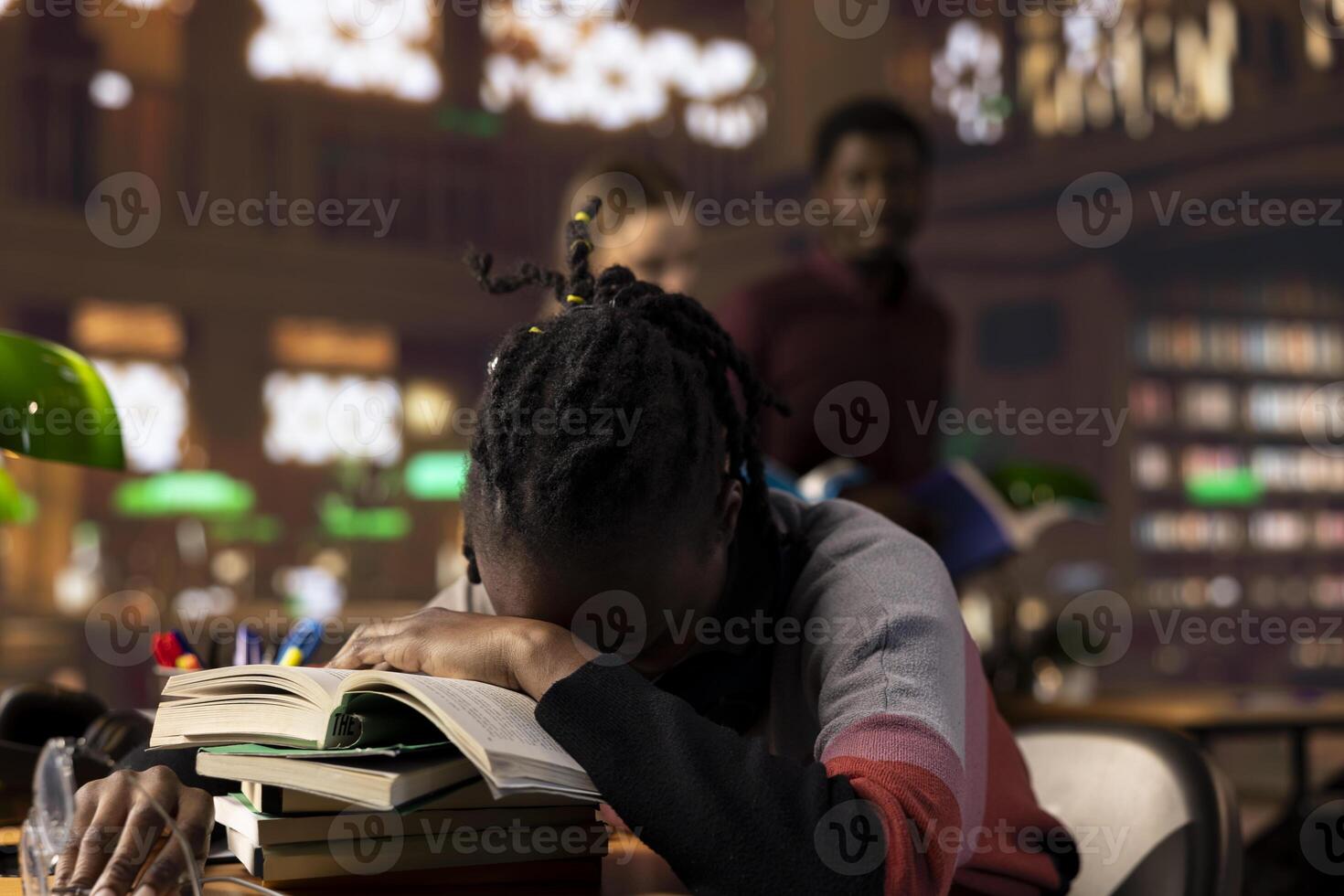 African american pupil sleeping on her books pile after a long night of studying, struggling to finish college and pass exams. Fatigued sleep deprived student resting in the library due to overload. photo