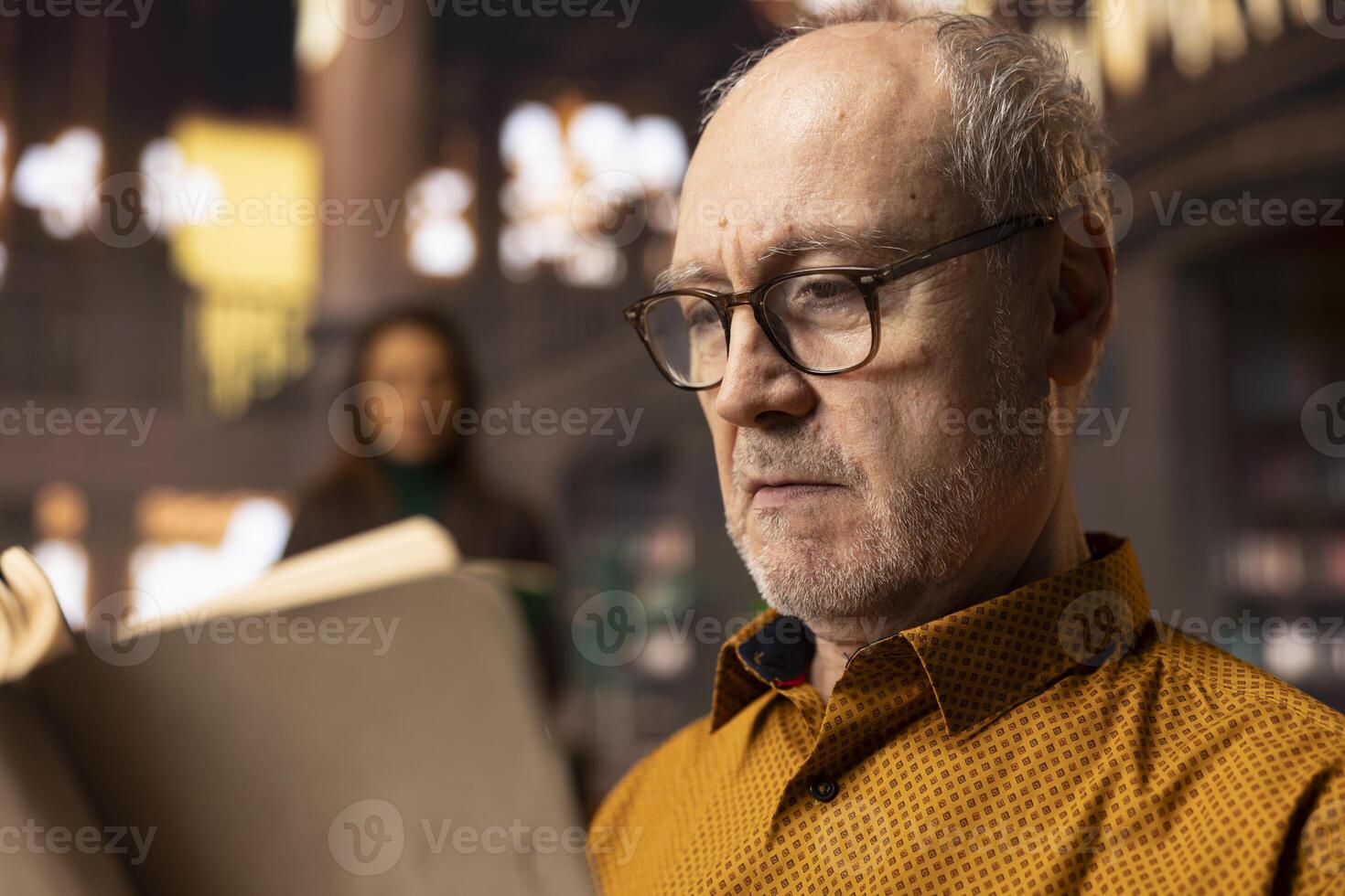 Retired man enjoys book reading at a public library and for leisure activity, feeling relaxed and increasing his knowledge during retirement. Happy calm person with eyeglasses, recreation. photo