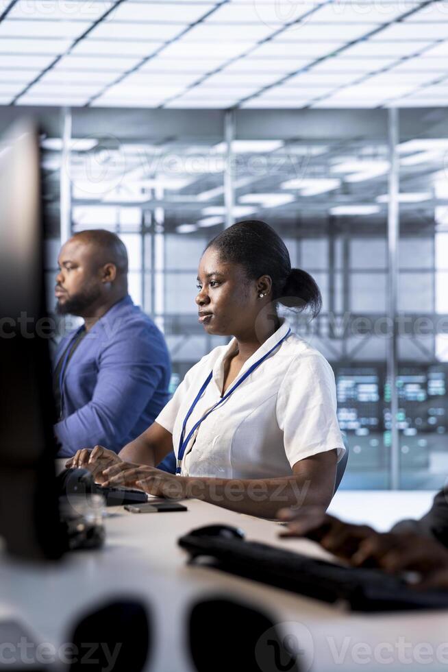 African american admin in data center using PC to check for server performance issues arising due to high CPU usage. Woman in server hub fixing insufficient memory errors slowing down hardware photo