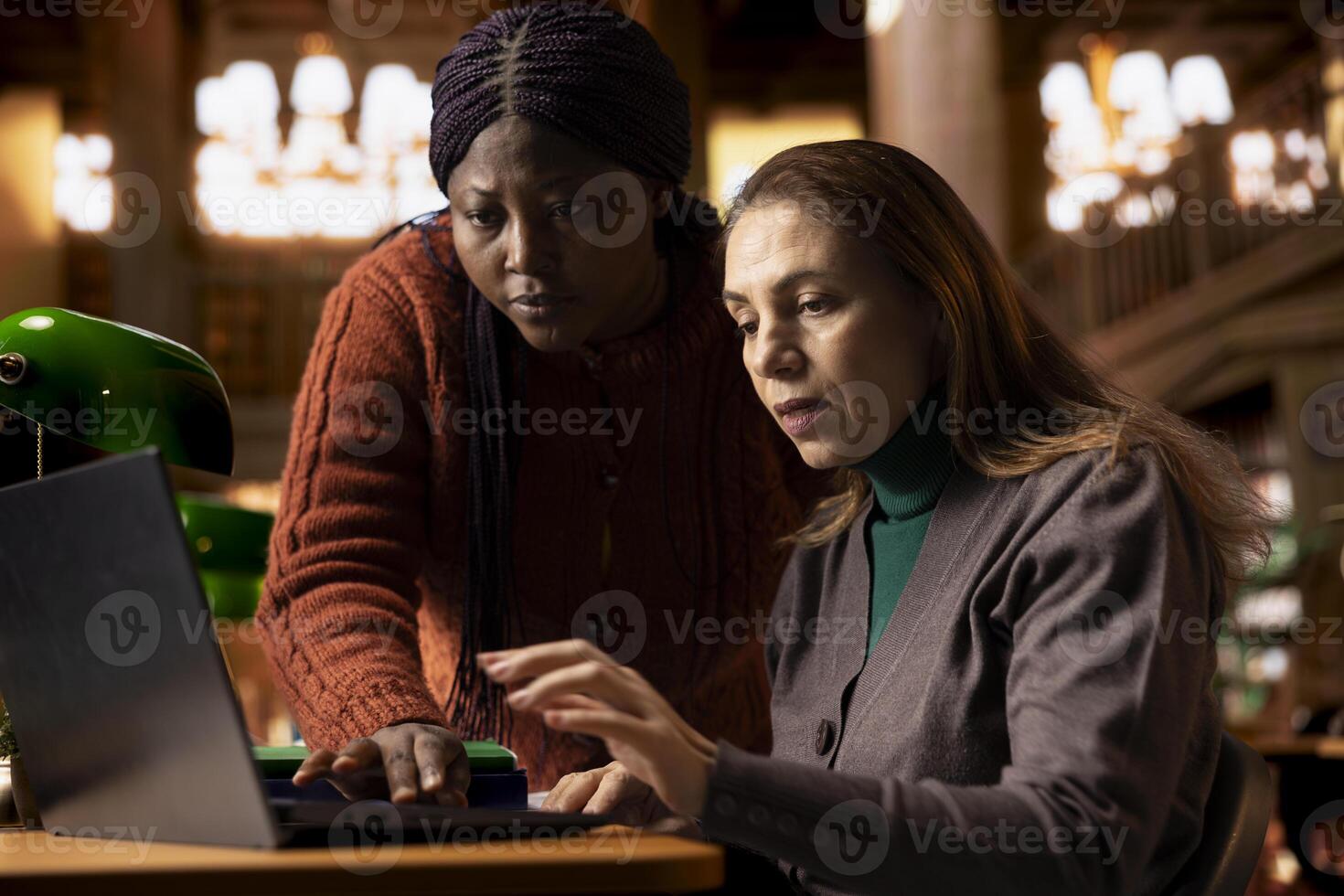 Team of diverse intellectual senior women reading a text book online, comparing class notes and sharing ideas with each other for a group project. Two colleagues discussion in the library. photo