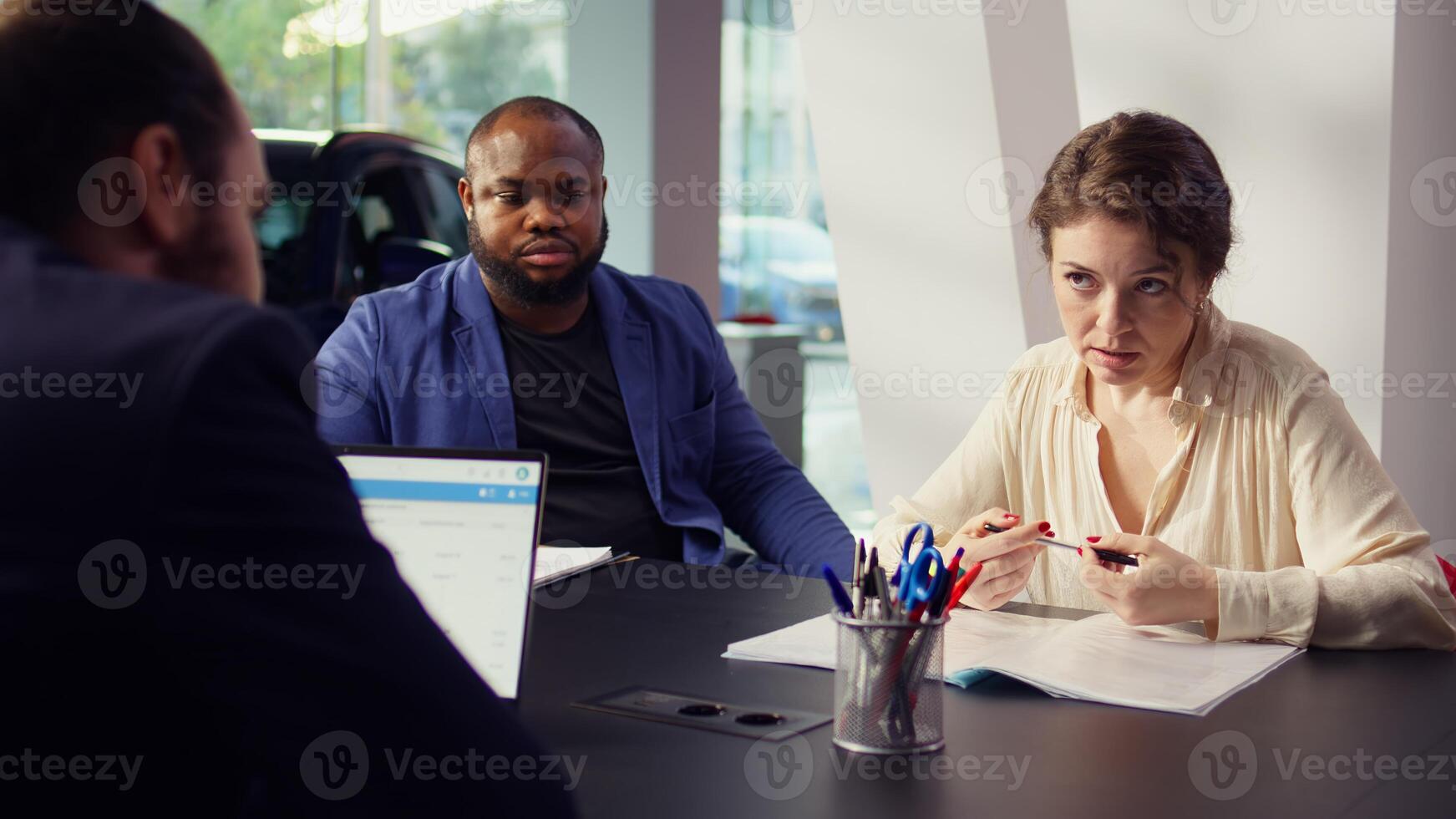 Client in car showroom deliberating purchase, negotiating with salesperson to reduce final price before deciding to sign contract. Woman in dealership debating whether to make vehicle sale, camera B photo