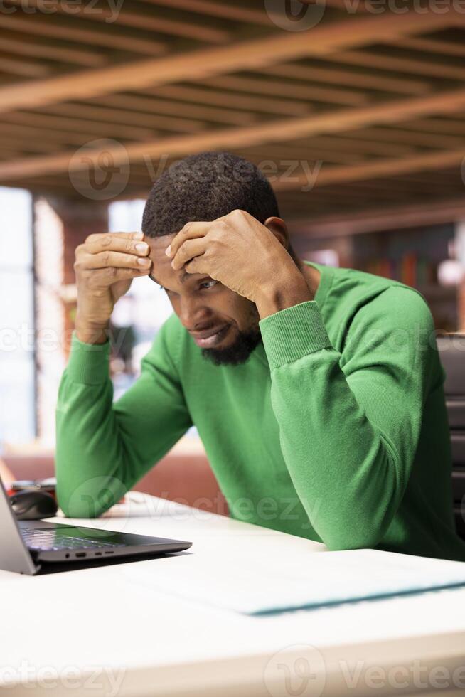 Man feeling clueless while preparing for school exam at home office desk, struggling to memorize info. African american student researching for college tests using laptop, doing brainstorming photo
