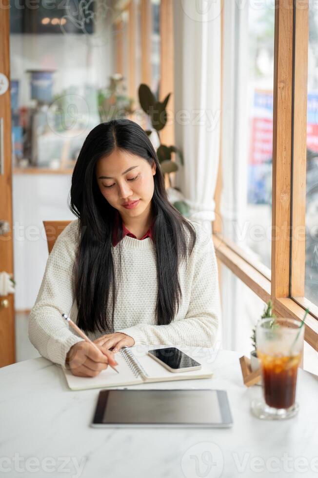 An asian woman is writing in the notebook, sitting with a tablet and a drink place on the cafe table photo