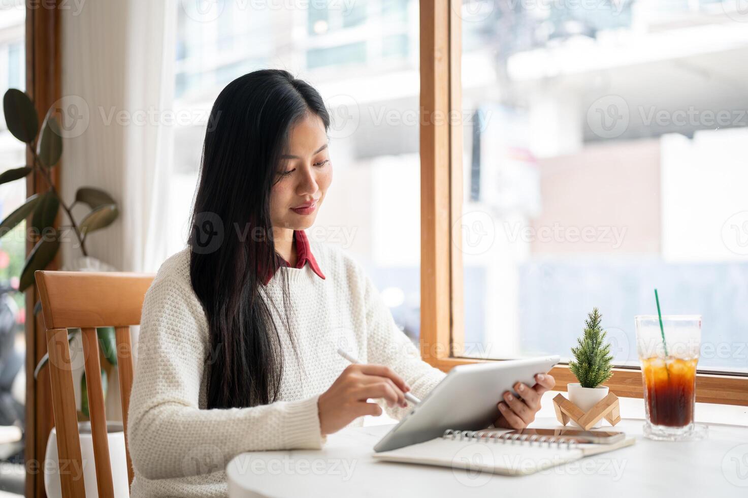 An asian woman is smiling while writing in tablet, sitting at the table beside large window walls. photo