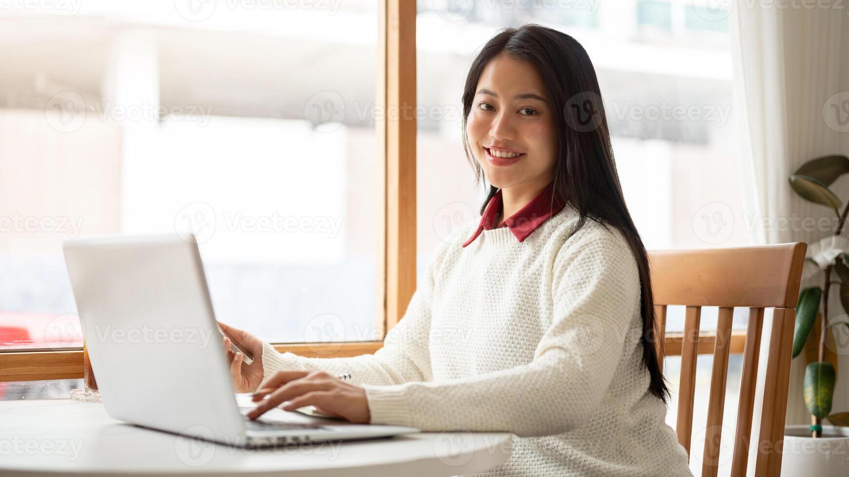 A smiling asian woman is typing on the laptop while holding the phone. Working or study in cafe. photo