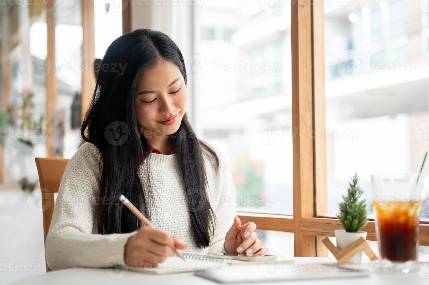 An asian woman is smiling while writing in the notebook, sitting at the table beside window walls. photo