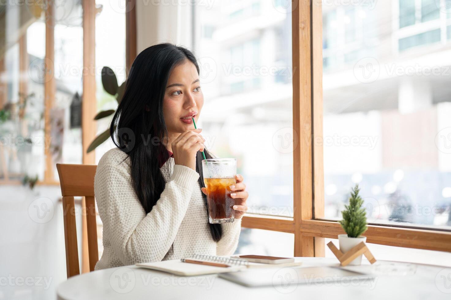 An asian woman is drinking the ice tea while looking through large window walls of the cafe. photo