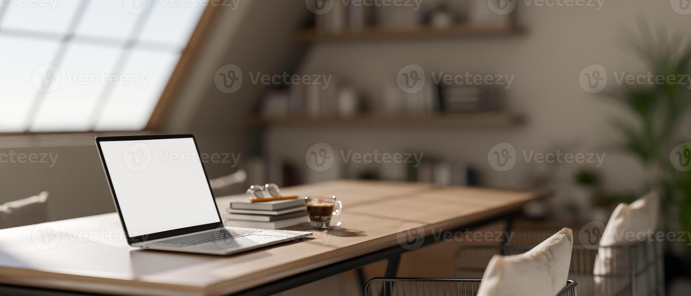 A white screen laptop on wooden table with books and coffee in a sunlit room room with large window. photo
