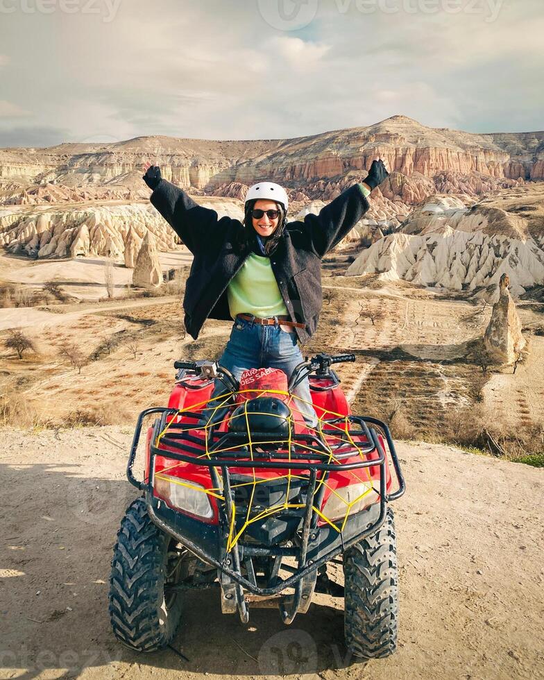Goreme, Turkey, 2024 -happy joyful excited woman adventurer hands up sit on red squad over Cappadocia red valley. ATV squad group tour viewpoint. Travel Turkey in winter outdoors photo