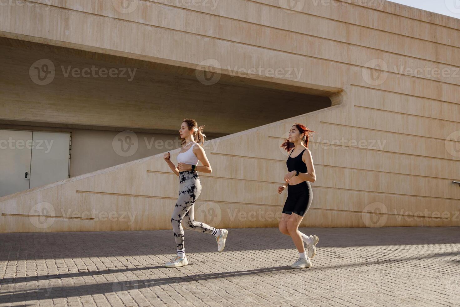 Two smiling women athlete running side by side along an outdoor track on modern buildings background photo