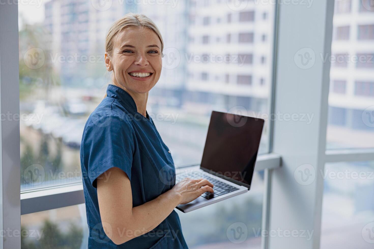 Smiling female healthcare worker using laptop while working at doctor's office and looking camera photo