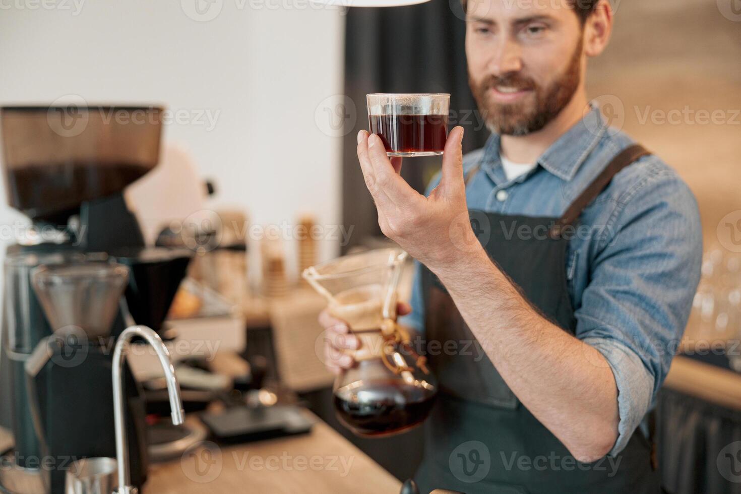 Close up of smiling barista appraisingly looks at a glass of filter coffee photo