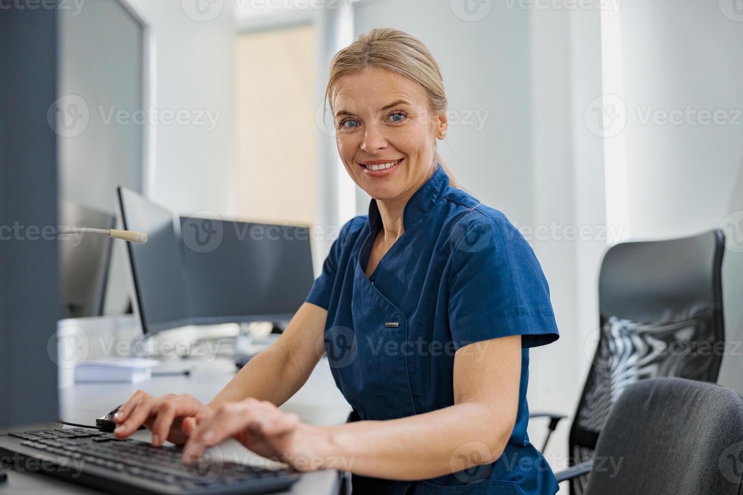 Nurse on Duty working on computer at the Reception Desk in modern clinic and looking camera photo