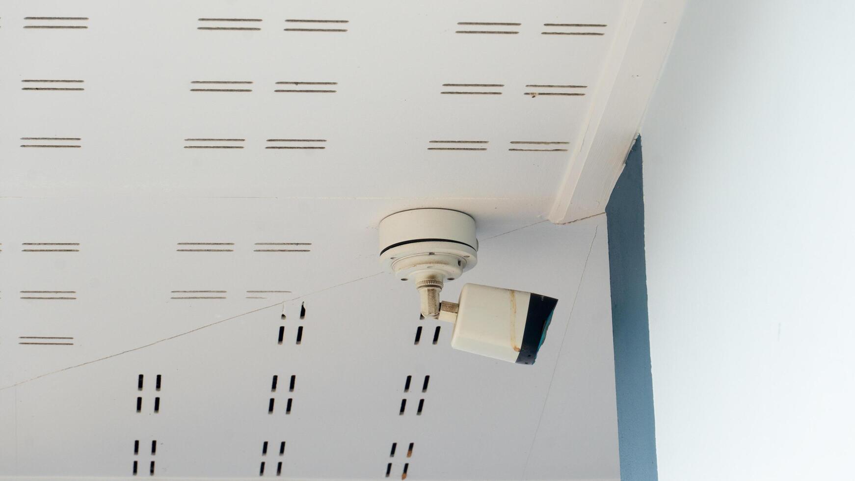 Outdoor CCTV camera. Installed under white ceiling panels. Camera looks towards the side of the building. Material is stained with what looks like rust. photo