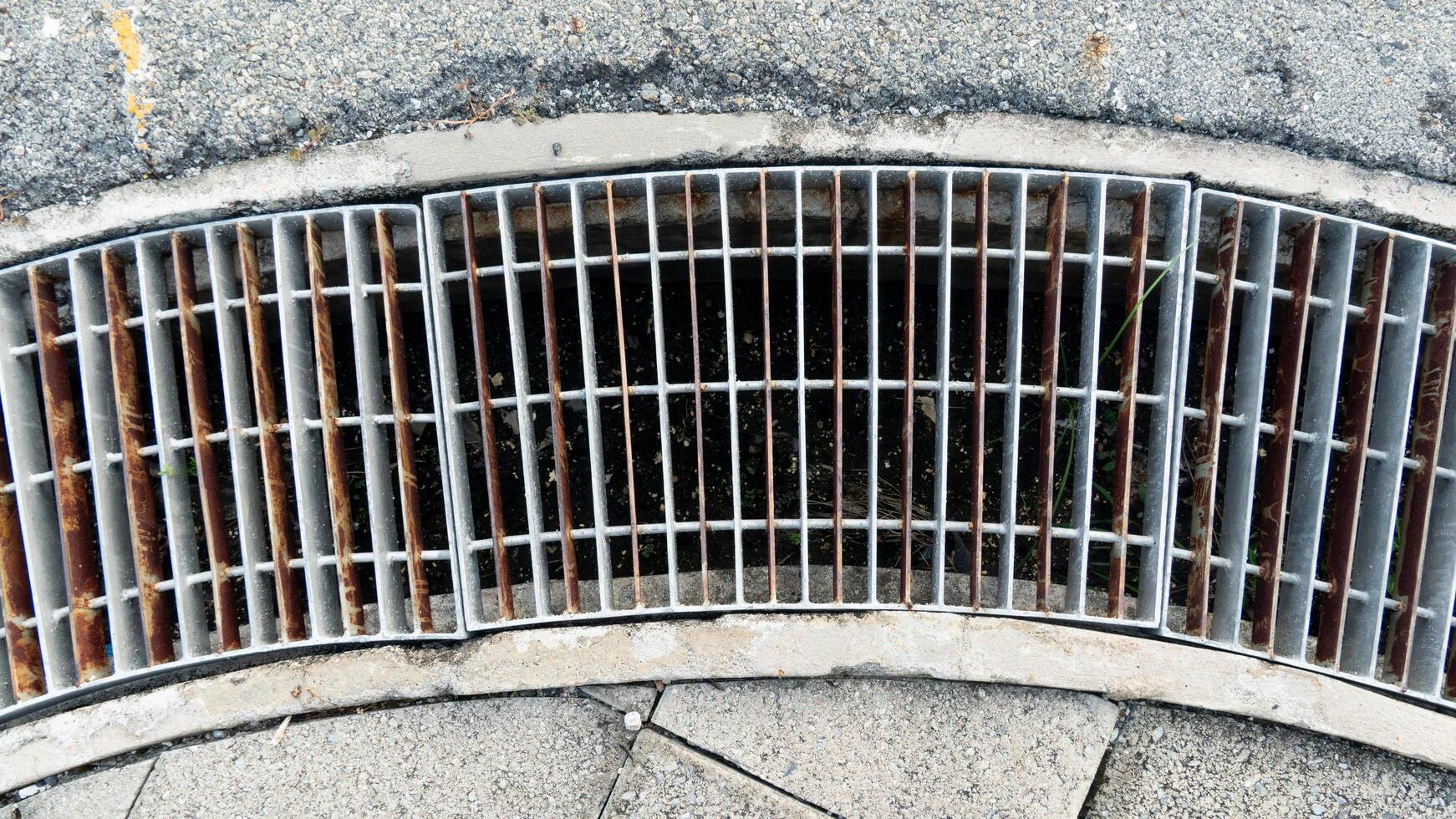 Curved drainage channel blocked by steel plates. Side of the footpath is made of concrete slabs. and asphalt road outside. photo