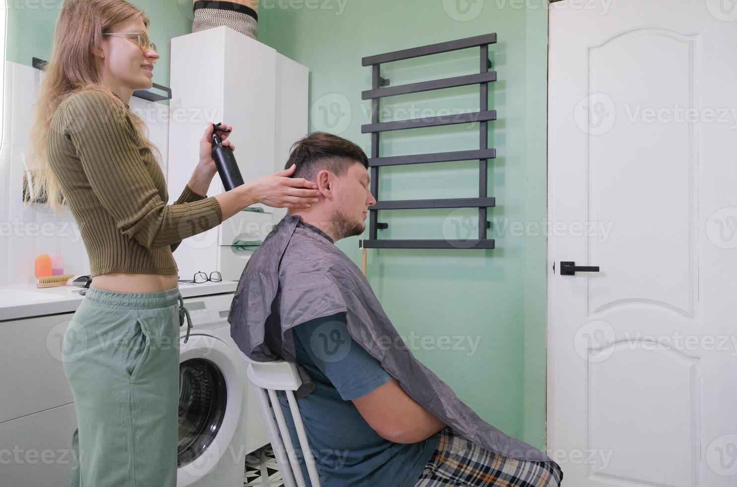 young caucasian woman cutting man hair in bathroom at home, spraying hair photo
