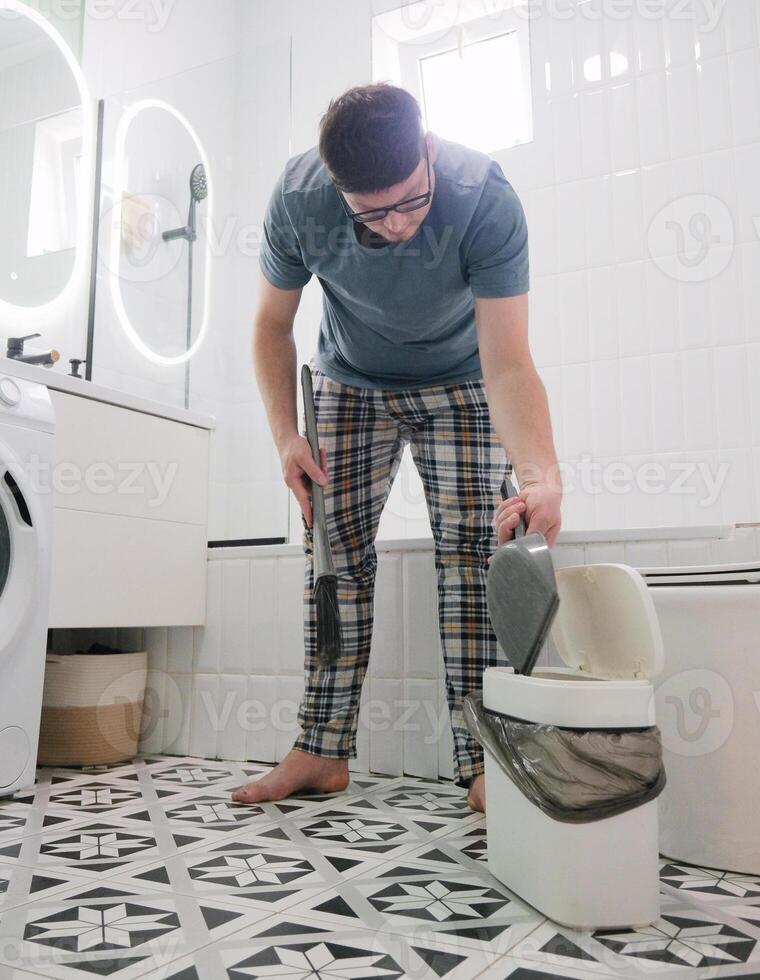 A man in home clothes sweeping floor in the bathroom, using garbage bin photo