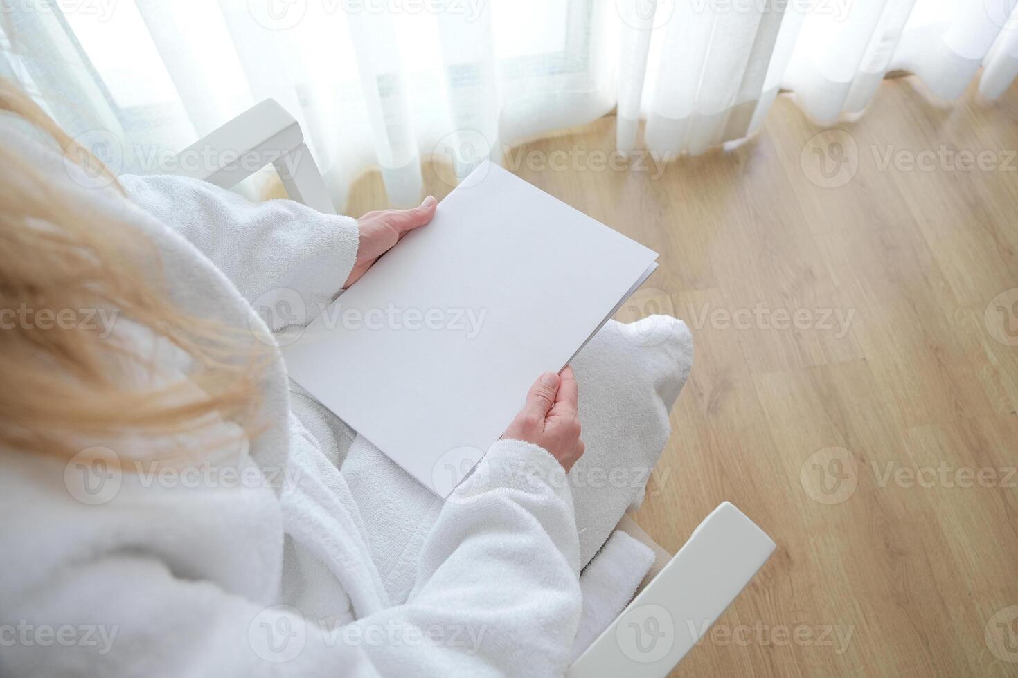 unrecognizable woman holding blank magazine for mockup design, sitting in chair in hotel room photo