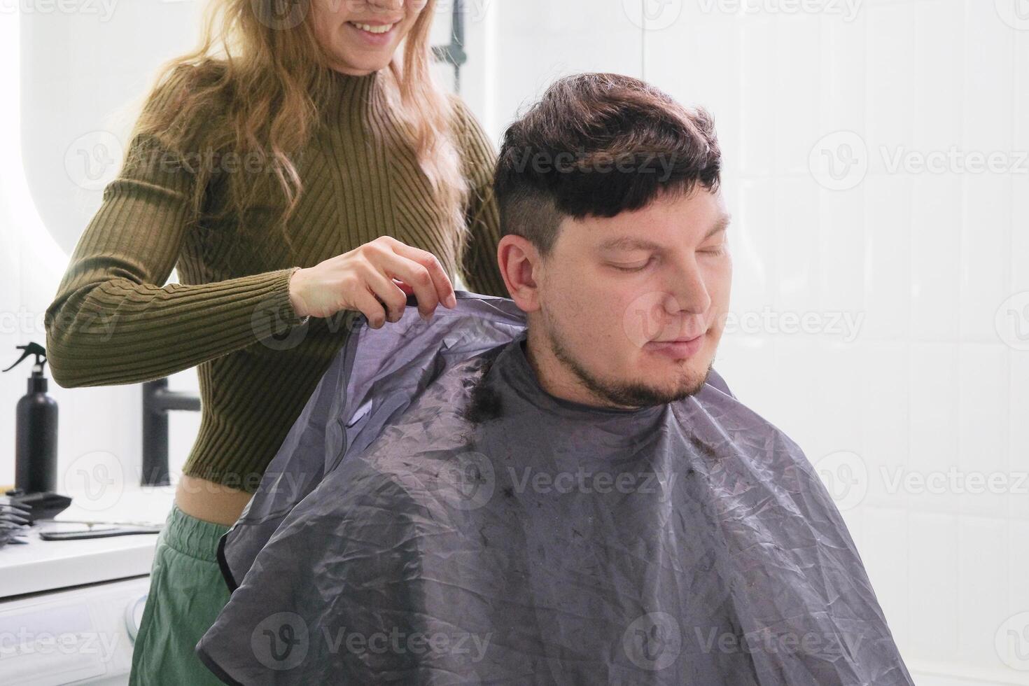 young caucasian woman cutting man hair in bathroom at home, taking off cover photo