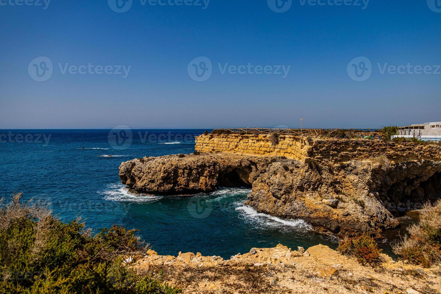 A rugged coastline with steep cliffs overlooks a calm, deep blue sea under a clear sky photo