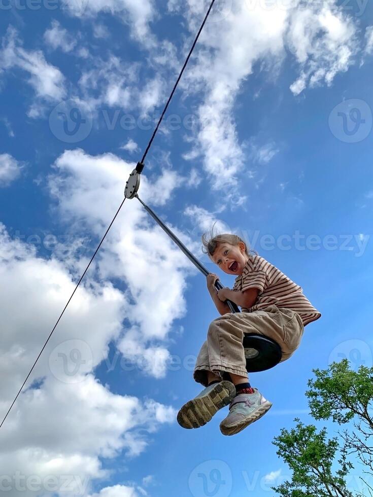 A happy cute little girl on a zip-line in summer. Bright blue sky and trees. photo