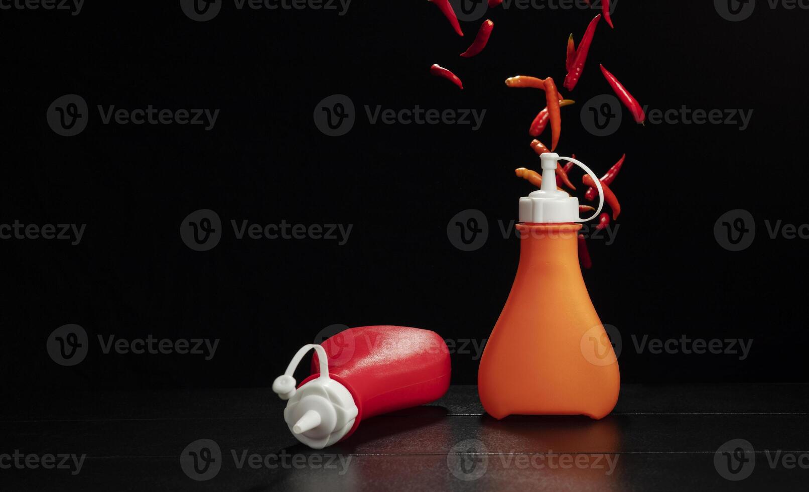 Bottles of chili sauce and ketchup are placed on the table with fresh red chili peppers, spicy food condiments, on a black and dark background. photo