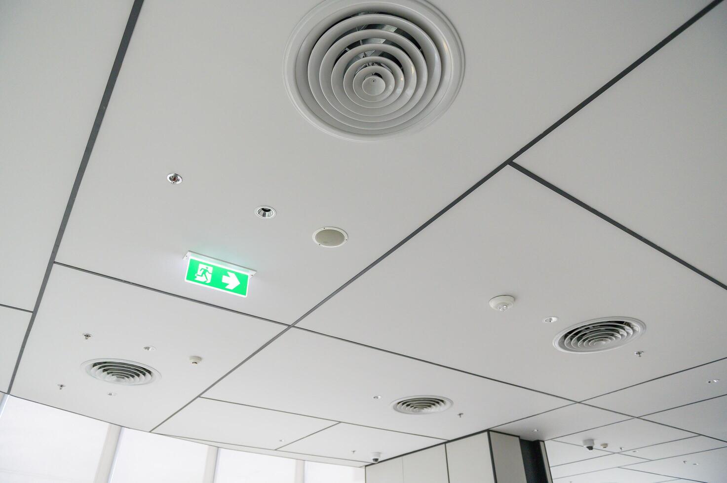 Clean modern office ceiling featuring air vents, recessed lighting, and grid pattern with emergency exit sign glowing green, creating bright and organized atmosphere photo