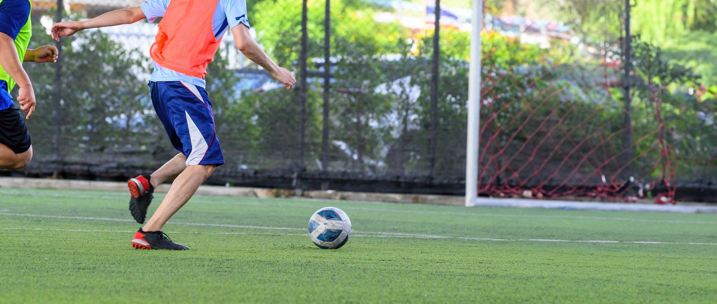 Soccer players in action on green grass field focusing on their legs and ball near goalpost during outdoor game with net and trees in background photo