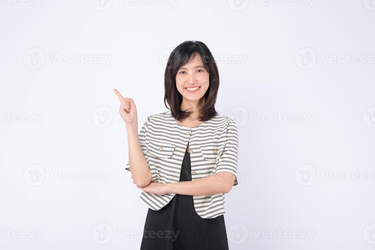 Cheerful southeast asian woman in striped jacket smiling brightly while advertising with her finger pointing up in clean white background photo