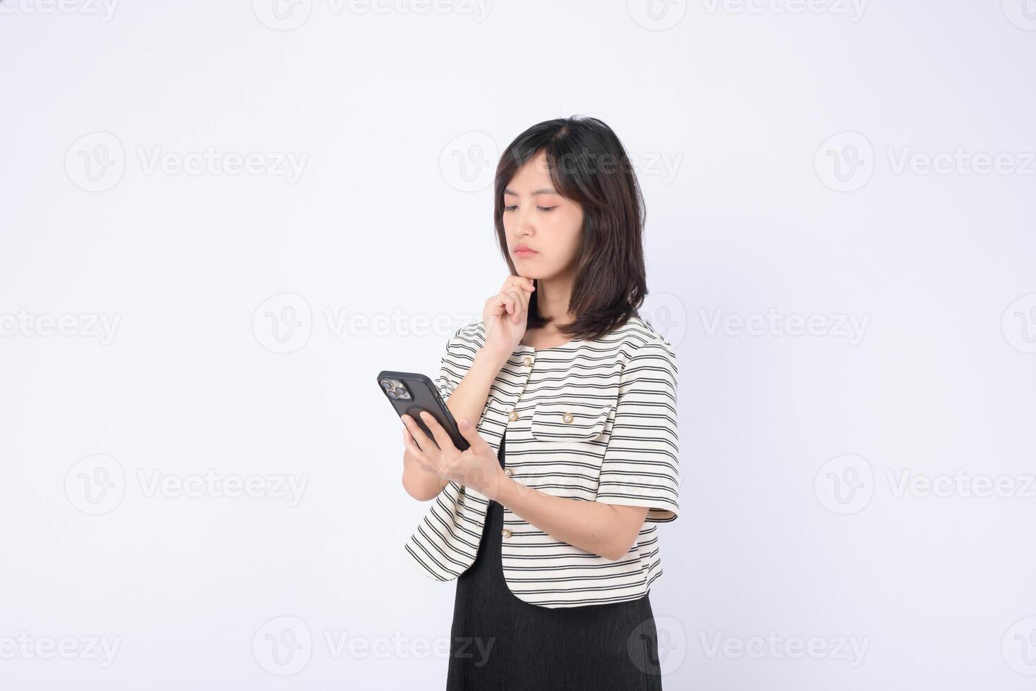 Southeast asian woman uses mobile phone with internet application looking thoughtful and focused on white background in casual striped jacket and black dress photo