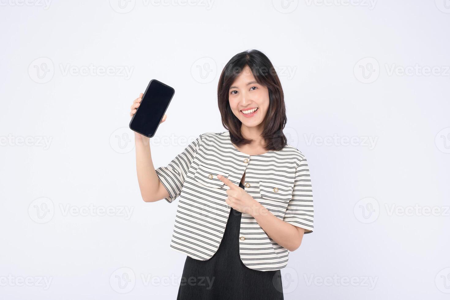 Southeast Asian woman smiling and pointing at mobile phone screen showing internet application in bright studio setting with casual striped jacket and black dress photo