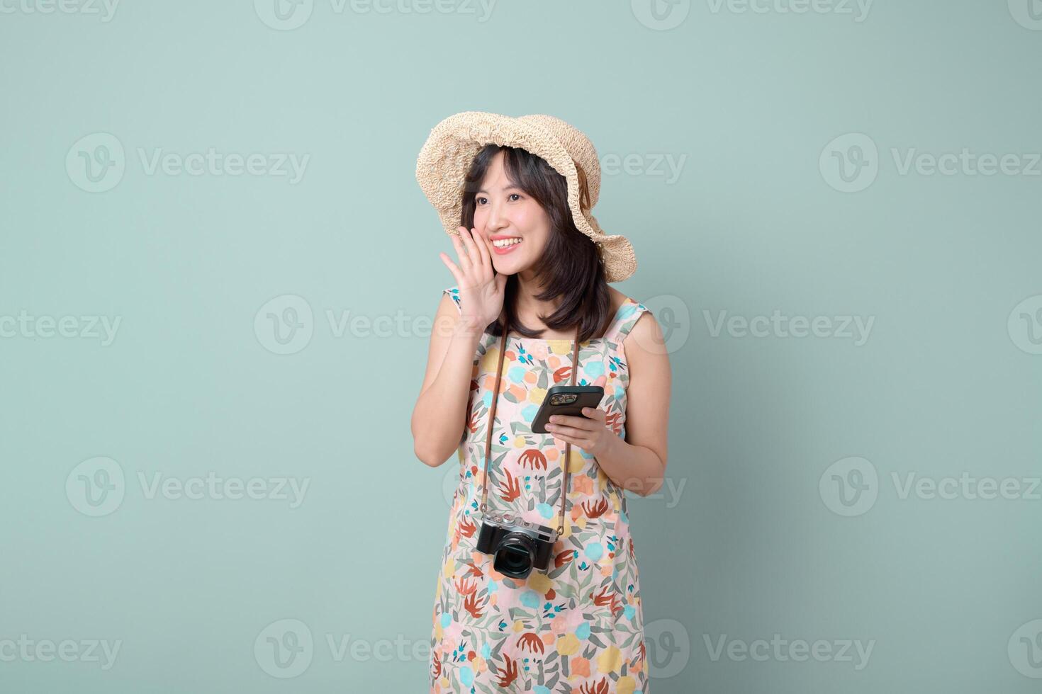 Southeast asian woman wearing floral dress and straw hat happily uses mobile phone with camera hanging around her neck while enjoying internet application on pastel background photo