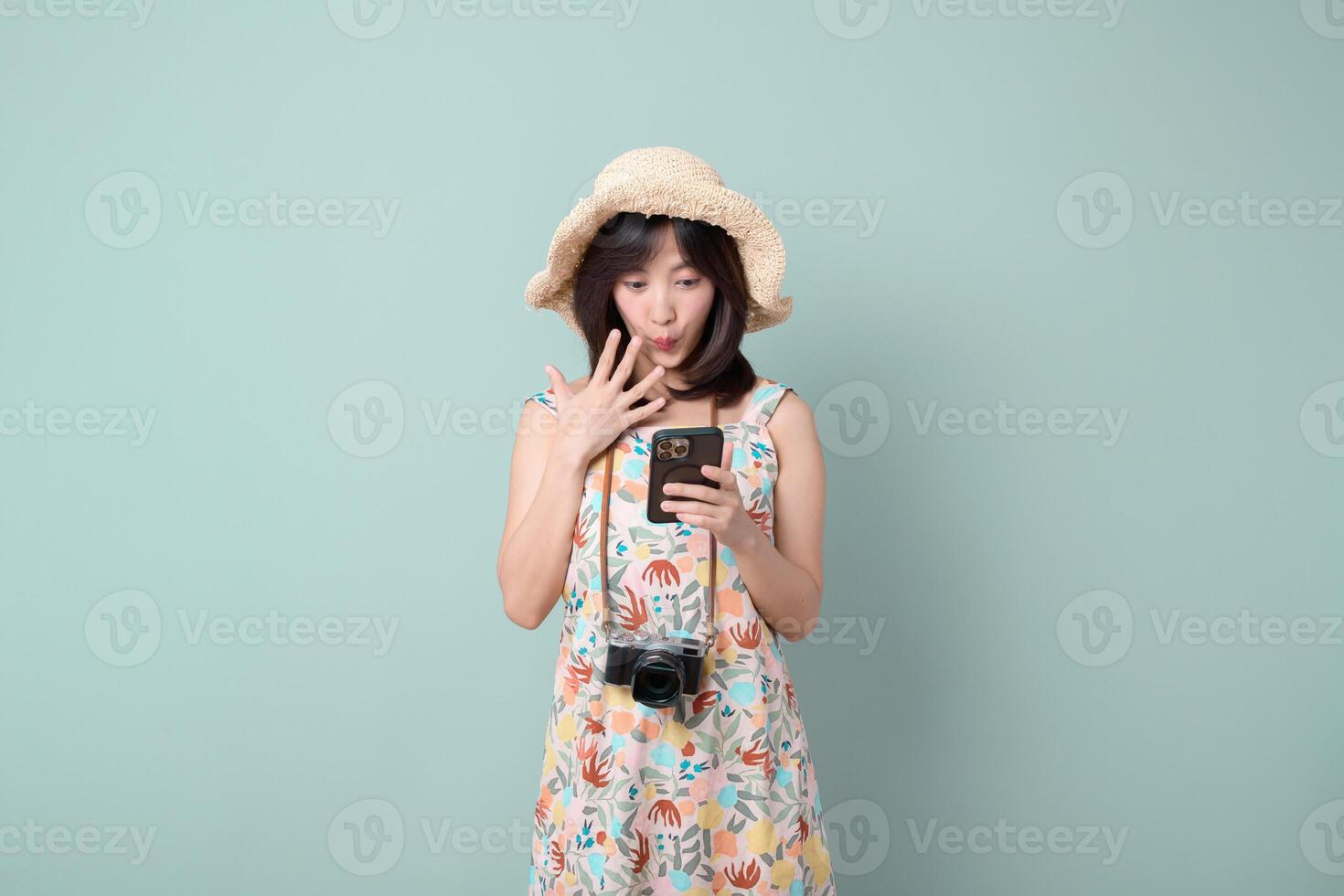 Southeast asian woman wearing floral dress and straw hat looks surprised while using mobile phone to browse internet application with camera hanging around her neck photo