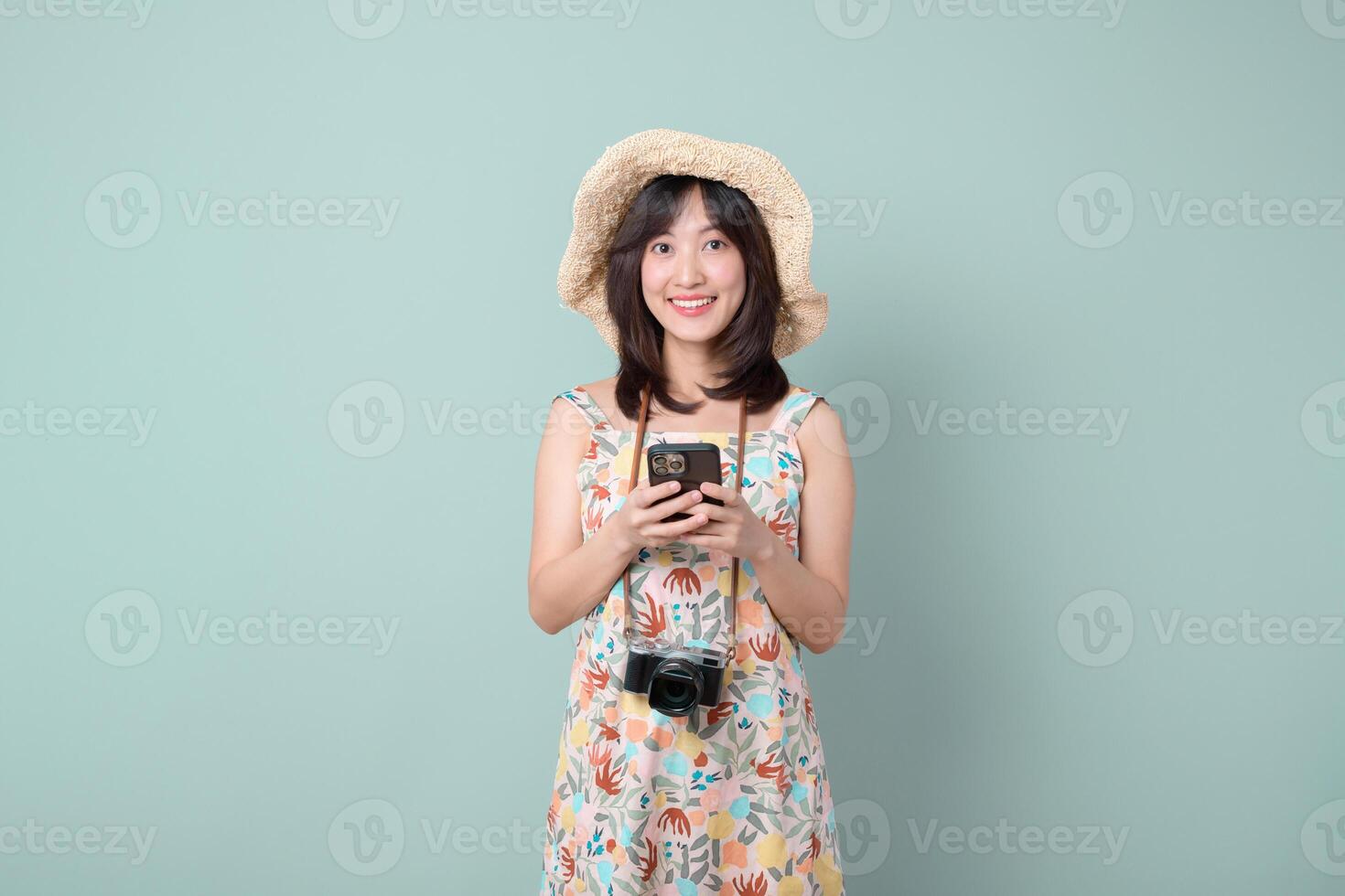 Southeast asian woman wearing floral dress and straw hat happily uses mobile phone with internet application while carrying camera around her neck in front of blue background photo