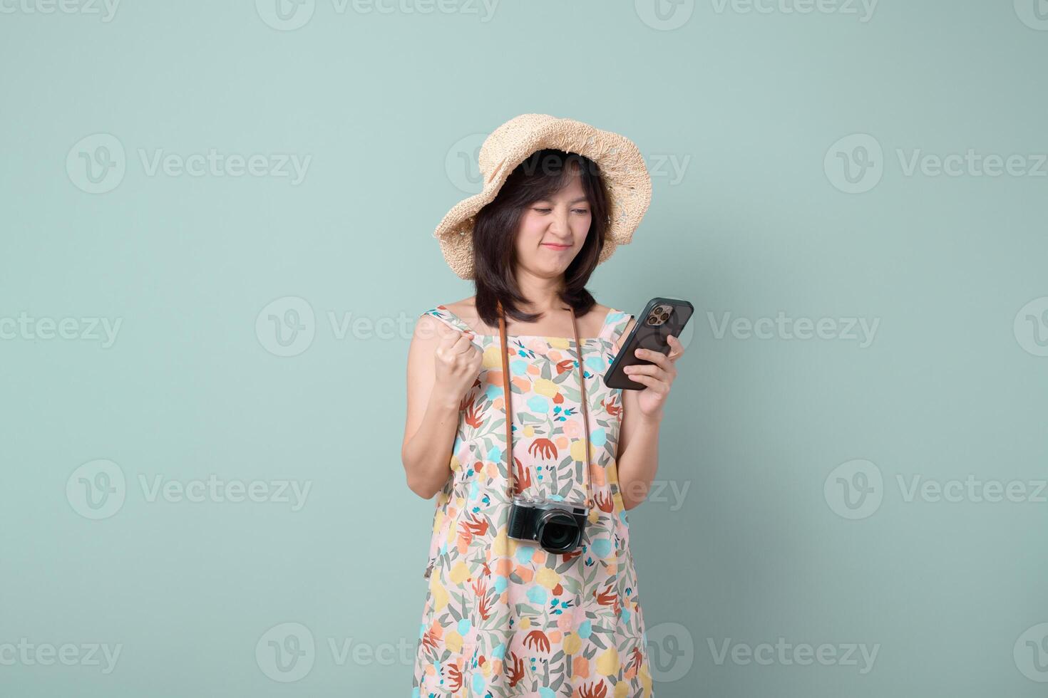 asian woman wearing floral dress and straw hat happily uses mobile phone to access internet application while holding camera around her neck in front of green background photo