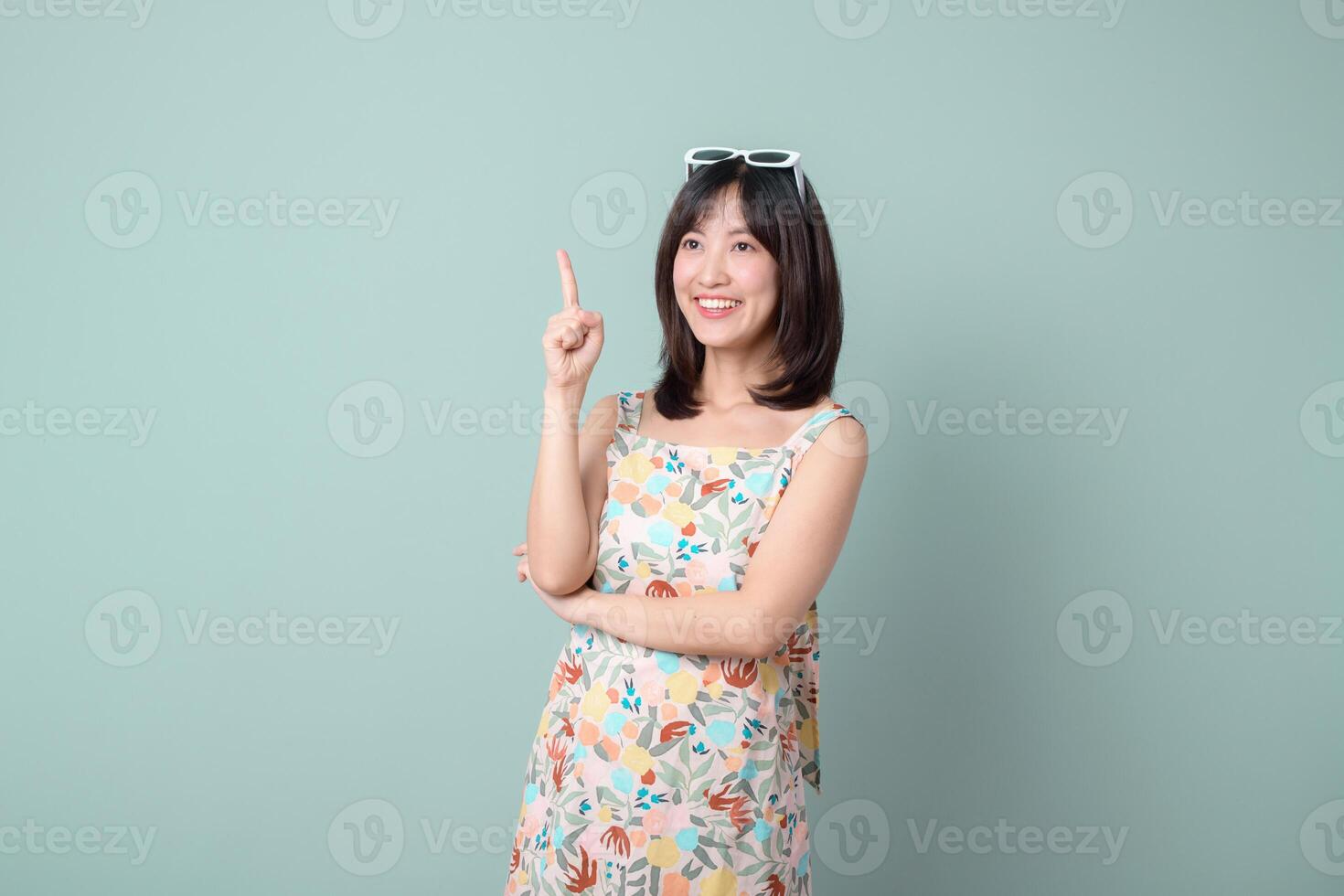 Southeast asian woman shopping online looks happy and confident while pointing upward wearing floral dress and sunglasses on her head against plain background photo