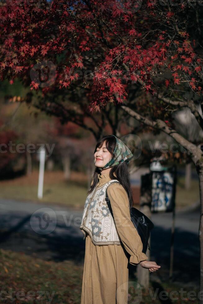 Southeast asian woman enjoys travel concept under red autumn leaves with closed eyes and peaceful smile wearing headscarf and beige dress in serene outdoor setting photo