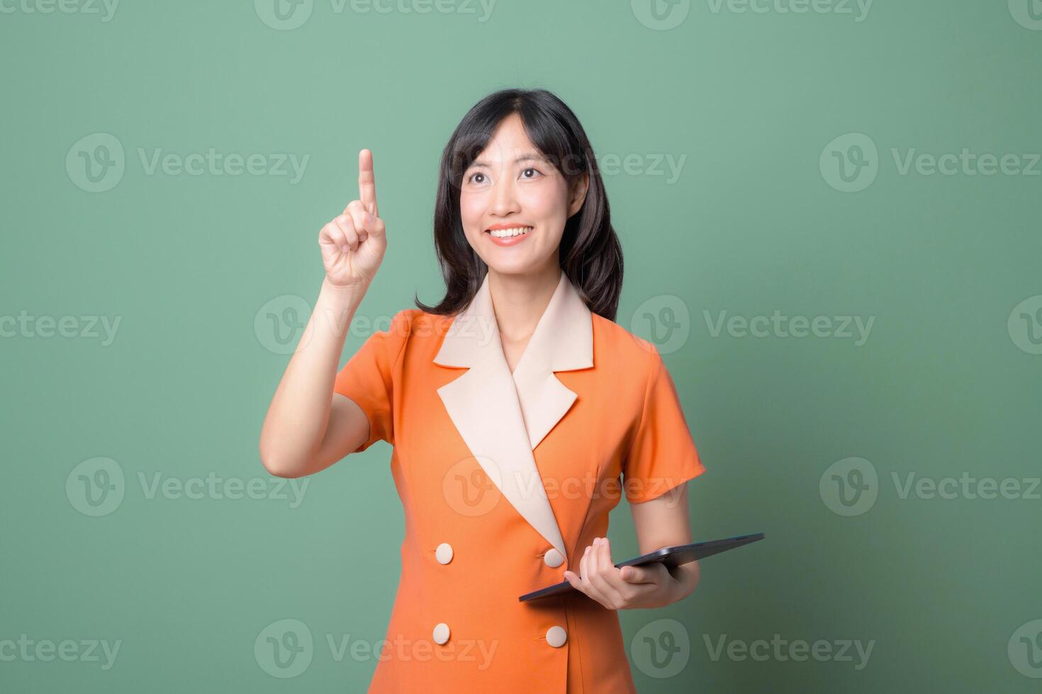 Southeast asian woman in orange suit smiling and pointing up while holding tablet against green background, expressing inspiration photo