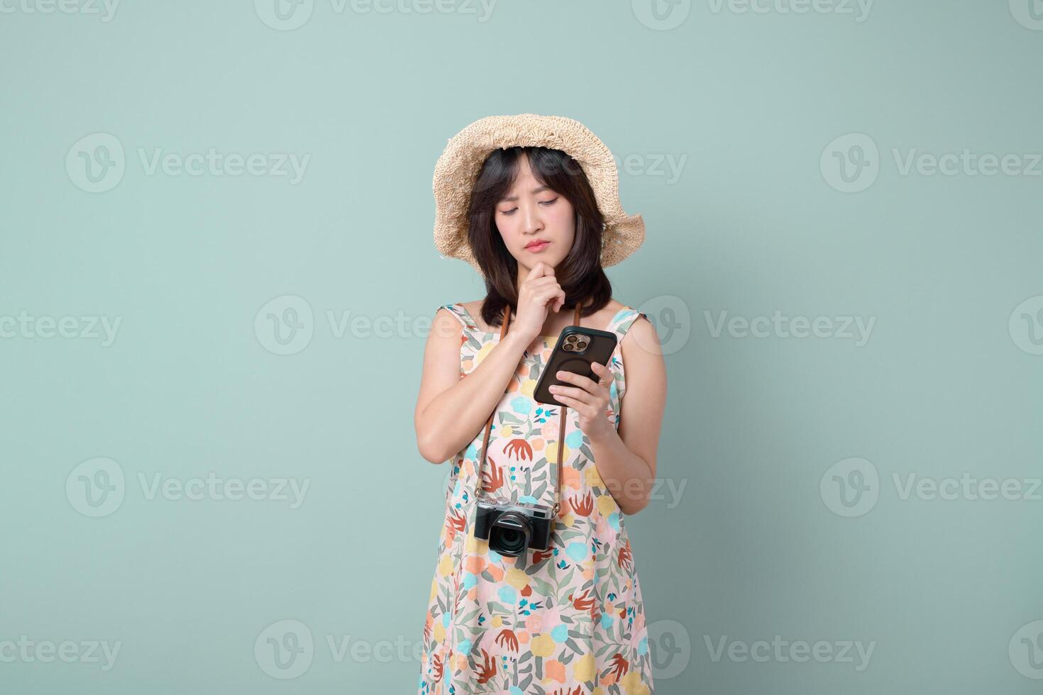 Thinking woman in summer dress and hat holding camera and mobile phone, standing on pastel green background, looking thoughtful photo