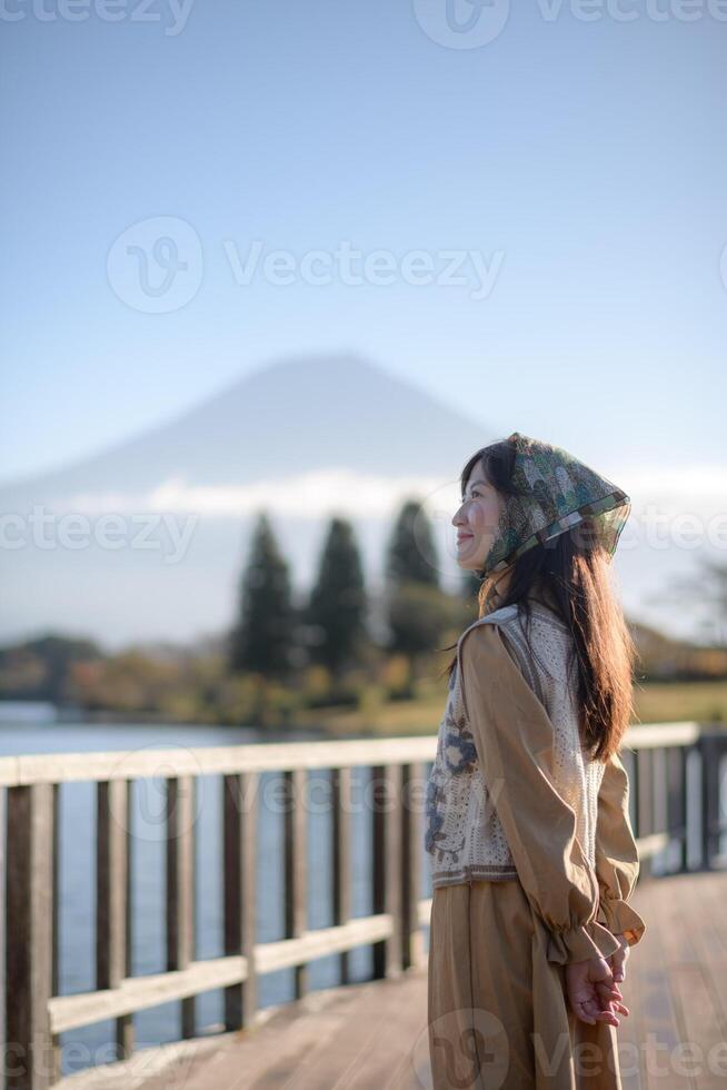 Southeast Asian woman stands on wooden deck near lake with mountain view in background, wearing patterned headscarf and beige outfit, enjoying peaceful outdoor scenery photo