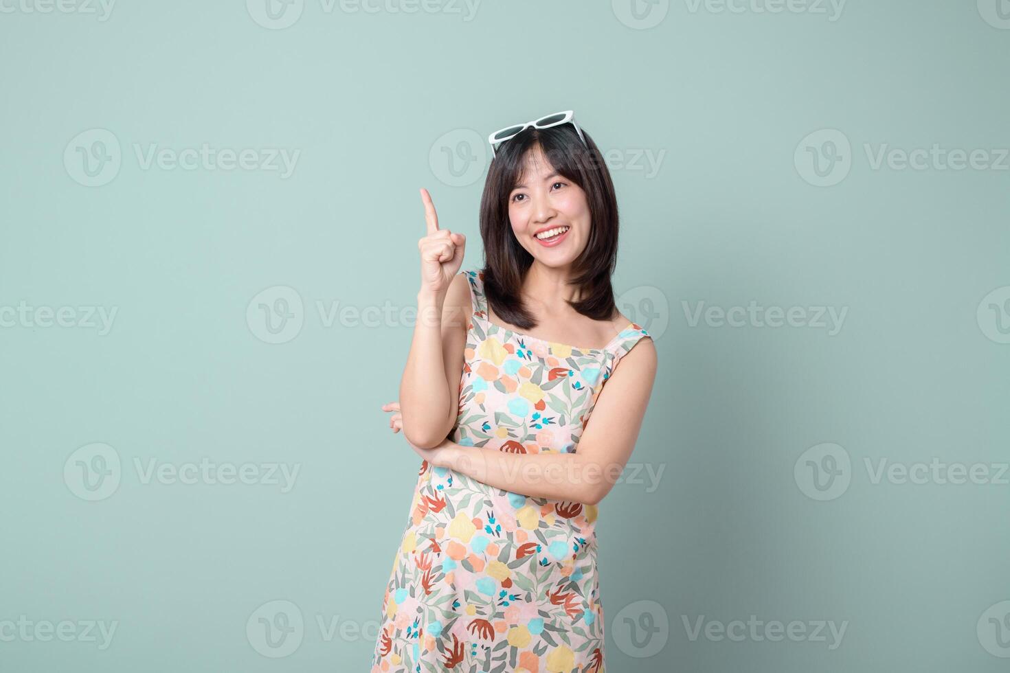 Southeast Asian woman in floral dress smiles and points upward with one finger against plain pastel background, expressing cheerful and positive mood photo