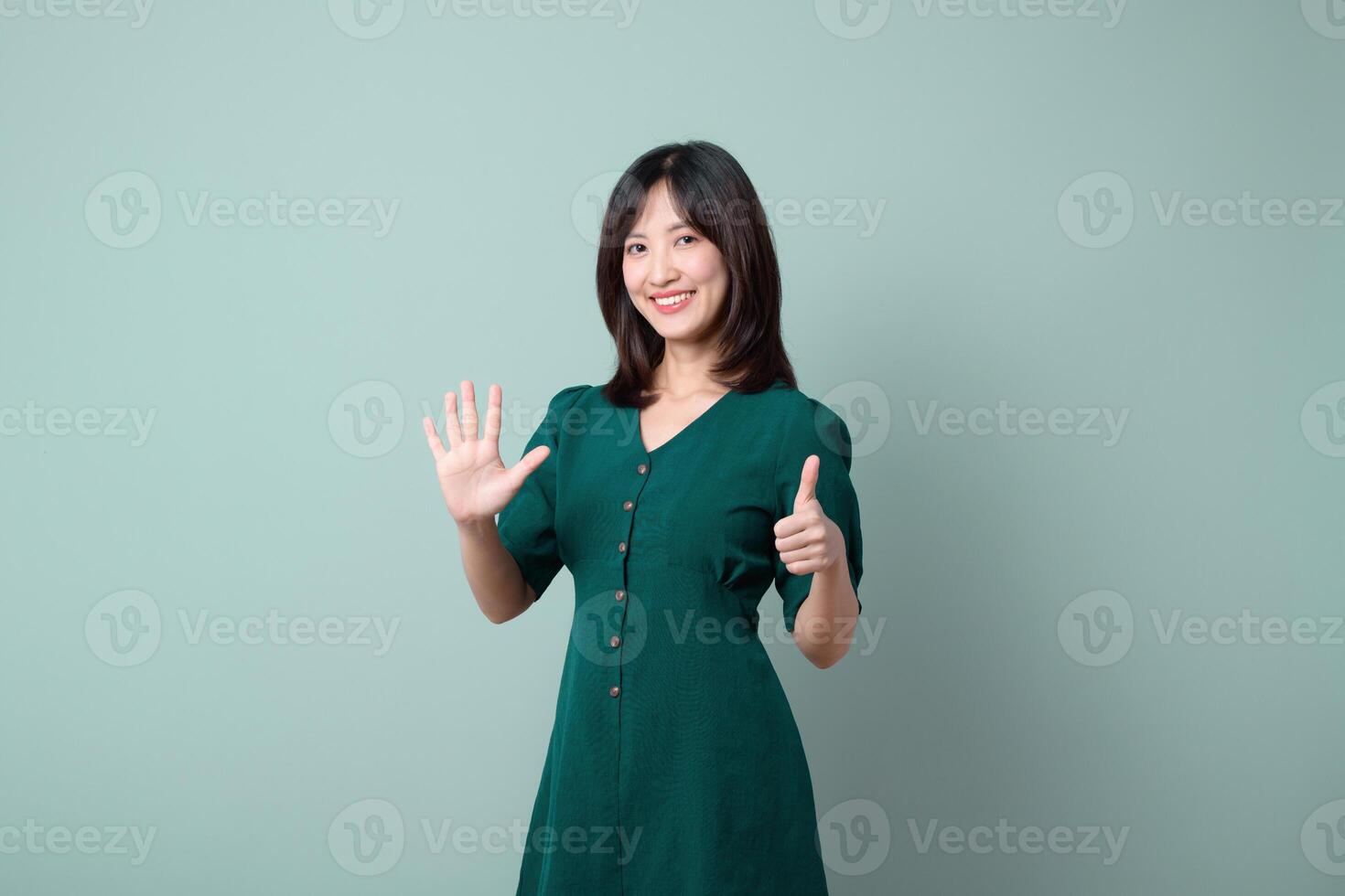 Southeast asian woman in green dress smiling and showing six fingers with one hand and thumbs up with other hand on plain background photo