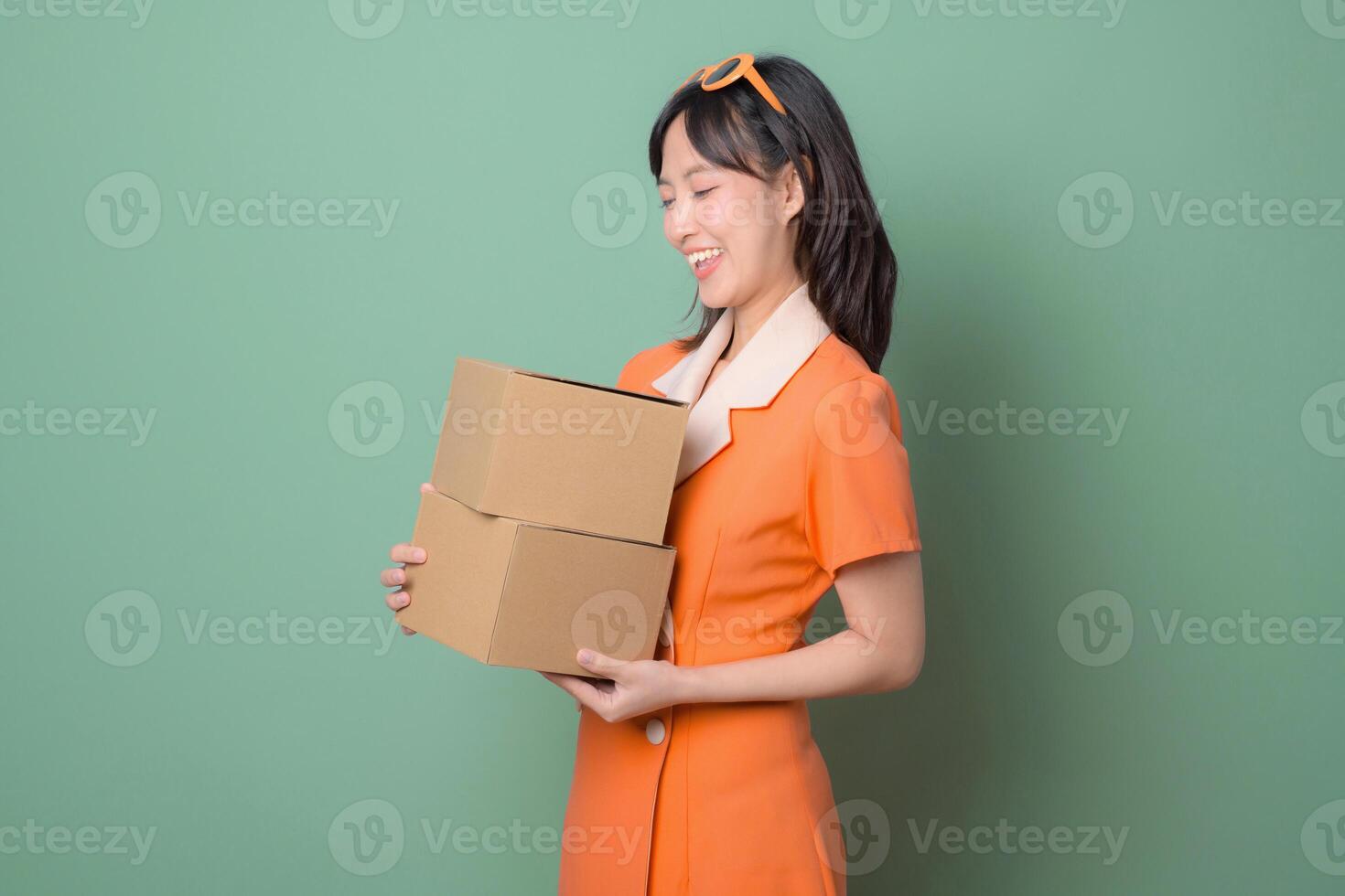Southeast asian woman in orange dress smiling while holding two cardboard boxes against green background photo